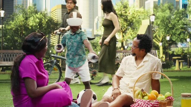 Boy wears a virtual reality headset and holds motion controllers while sitting between his parents on a blanket in an urban park, showing immersive VR technology during family picnic fun.