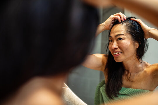 Smiling middle-aged asian woman checking hair in bathroom mirror