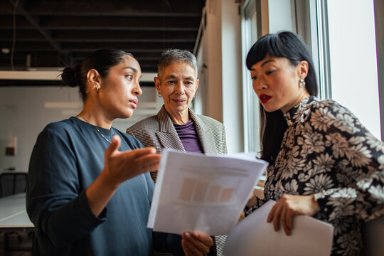 Three businesswomen discussing reports by a window in a modern office