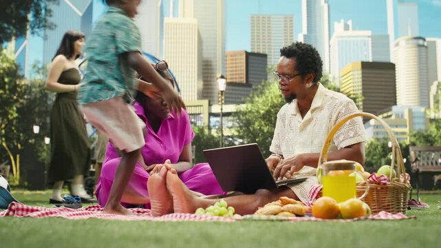 Workaholic father ignores his family while doing digital work outdoors as a concerned mother asks him to join their boy in playing on the picnic blanket.