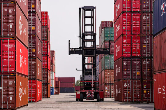 Worker with a tablet inspects rows of stacked shipping containers at a busy import export freight terminal yard outdoors.