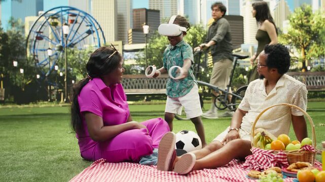 Boy wears a virtual reality headset and holds motion controllers while sitting between his parents on a blanket in an urban park during a family picnic.