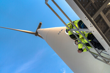 Rope access technicians in safety harnesses inspect the blade root from the nacelle deck seen from directly below on a wind turbine nacelle in clear blue daylight at a wind power plant in. © ultramansk