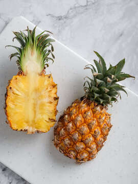 Two pineapples whole and halved on white speckled cutting board