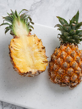 Two pineapples whole and halved on white speckled cutting board