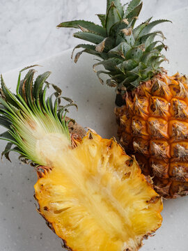 Two pineapples whole and halved on white speckled cutting board