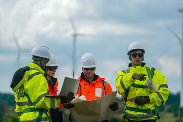 Fototapeta premium A mixed team of wind farm engineers in yellow and orange hi vis walk the meadow while consulting the site plan on a green meadow with rows of turbines behind in bright overcast daylight at.