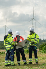 Fototapeta premium A mixed team of wind farm engineers in yellow and orange hi vis debate cable routing over a large site plan on a green meadow with rows of turbines behind in bright overcast daylight on a.