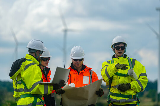 A mixed team of wind farm engineers in yellow and orange hi vis walk the meadow while consulting the site plan on a green meadow with rows of turbines behind in bright overcast daylight at.