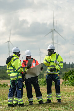 A mixed team of wind farm engineers in yellow and orange hi vis debate cable routing over a large site plan on a green meadow with rows of turbines behind in bright overcast daylight on a.