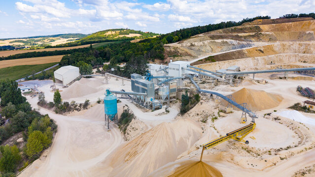 Conveyor machine dropping washed sand onto a large pile. Blue silo and sorting machinery operate near sand piles with surrounding green agricultural fields.