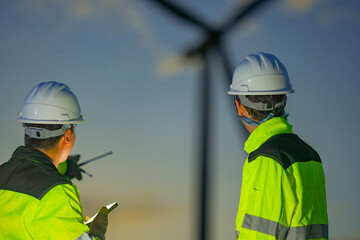 Fototapeta premium Two wind turbine technicians look up at the rotor while talking on the radio beneath the silhouetted rotor of a turbine in pastel dusk deep inside a tropical wind energy park.