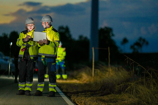 A wind energy team on the park road walk home along the service road with a glowing laptop on an asphalt road crossing the wind farm in black night at a wind power plant in Southeast Asia.
