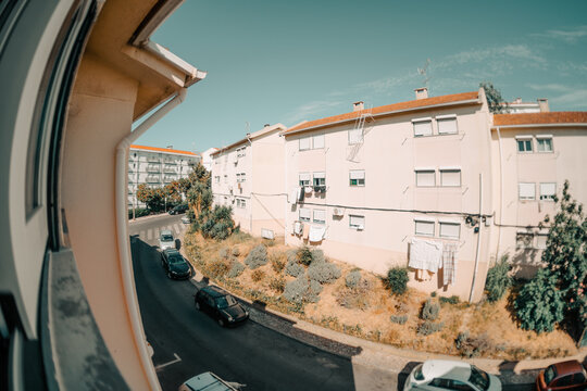 Wide distorted view from an open window overlooks modest apartment blocks, parked cars, hanging laundry and a dry landscaped courtyard beneath a clear blue sky