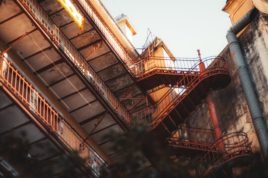 Ornate red iron fire escape twists between retro weathered gallery balconies in an aging inner courtyard, viewed upward as warm sunlight reveals peeling concrete and industrial character