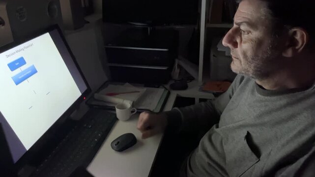 Mature professional man working on a decision-making flowchart on his computer and scrolling his smartphone in a dark office illuminated by the monitor's light