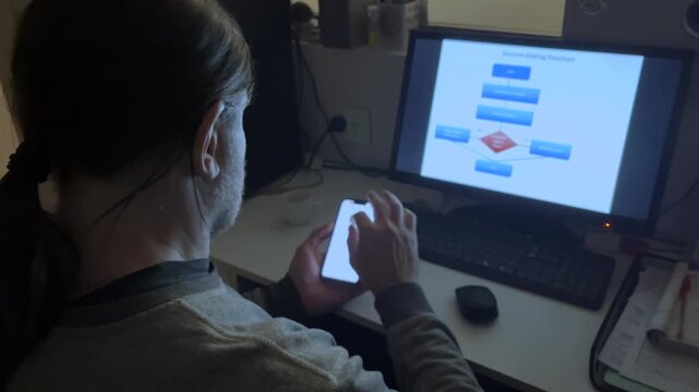 Focused male data analyst working in a dim office using a smartphone while a decision-making flowchart is displayed on the computer monitor in front of him