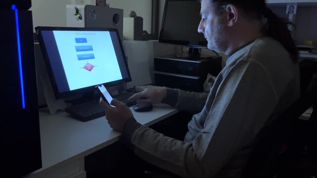 Concentrated man sitting at his desk at night, illuminated by the displays of his smartphone and monitor. Horizontal real time video