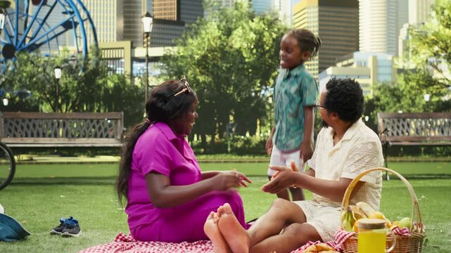 Young boy dances and has fun with his parents on a checkered blanket, fooling around and laughing with silly moves during a picnic and family leisure beside a fruit basket in an urban park.