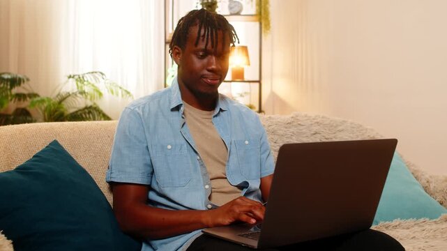 African American young man on home sofa works on laptop checking text and fixing small details with focused attention. Black guy on home sofa edits a document and reviews work with calm concentration.