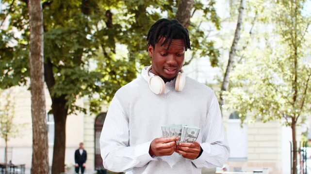 African American young man counting cash walking in city street after payment in park. Black guy tourist smiling outdoors while checking money with happy face during leisure break and travel success.
