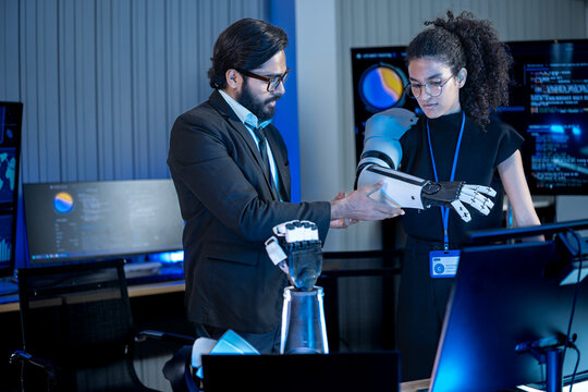South Asian male developer looks at a robotic hand demonstration by a Black female technologist in a dark AI research lab.