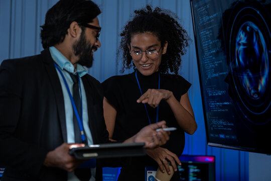 South Asian male engineer and Black female developer examine AI-driven code on a large screen in a blue-lit tech operations room.