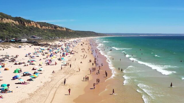 People gather on the beach under the sun aerial view. Children play in the sand while others relax on towels. Colorful umbrellas dot the area as waves roll in.