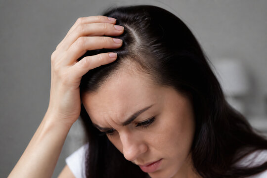 A woman with dark hair is touching her scalp in a distressed manner indoors.