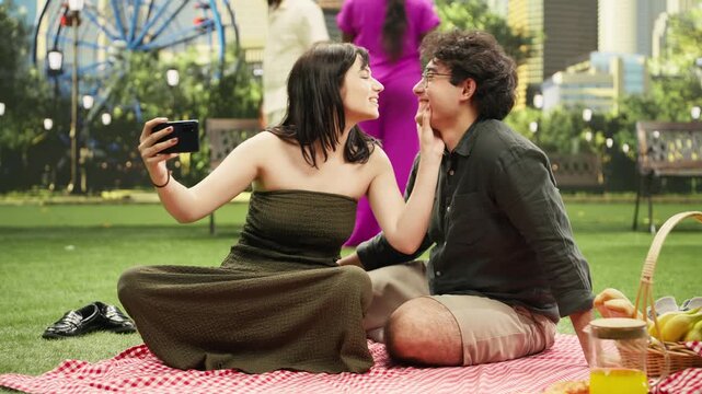Young couple take playful photos on a mobile phone during a picnic date in an urban park, smiling and bonding on a blanket while enjoying a romantic date with smartphone selfies together.
