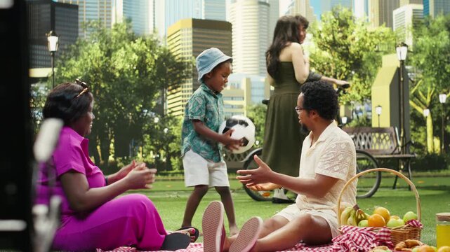 Parents and boy play catch with a soccer ball beside a picnic basket and blanket in an urban park while enjoying an active family day together.