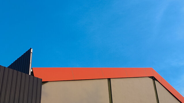 Minimalist architectural detail of a modern building rooftop against a clear blue sky. Features a bright red-orange roof edge, beige wall panels, and dark corrugated metal siding. 