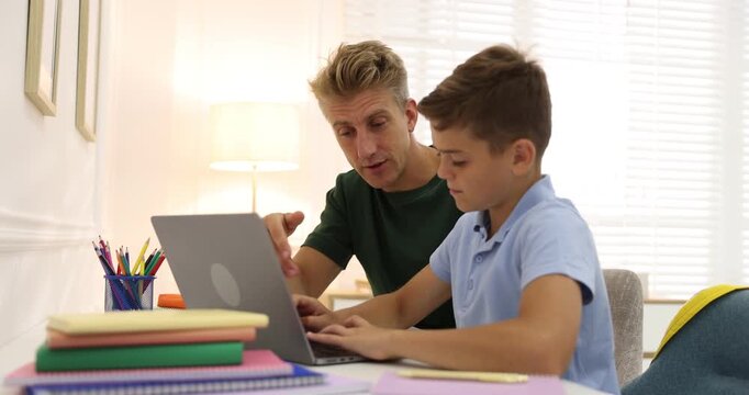 Father helping his son with homework at table with laptop indoors