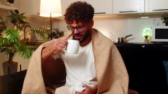 Arabian man at home feels sore throat holds mug of hot tea with medicine takes slow sip uses tissue. Egyptian guy on sofa searches doctor on smartphone rubs neck coughs rests under blanket sniff fever