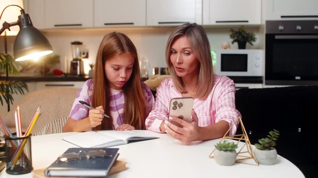 Mother and daughter watch math tutorial on smartphone parent explains example child writes notes in notebook stays focused and learns together. Woman with teenager girl sitting at table at evening.