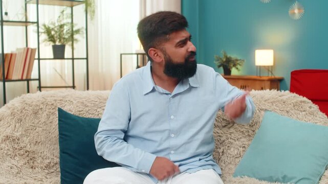 Indian man at home strums invisible guitar feels rhythm imagines rock stage laughs enjoying great music. Arabian guy on sofa waves arms energetic fun performance shows playful spirit and creative joy