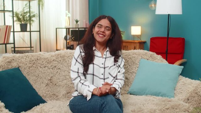 Indian woman at home laughs heartily eyes radiating happiness joy positive stress relief vibe. Arabian girl on sofa chuckles sincerely claps hands showing amusement fun carefree mood playful spirit.