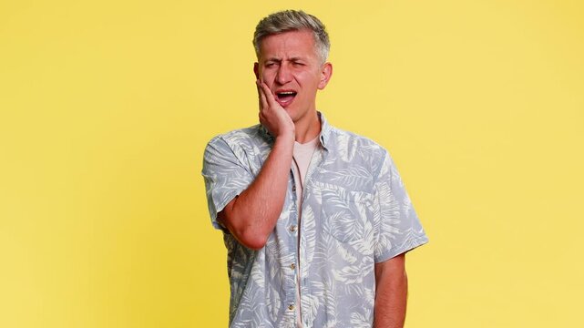 Middle-aged man holding cheek with tense painful face, grimacing from sharp toothache or dental problem, clearly suffering. Young guy showing health pain expression isolated on yellow background.