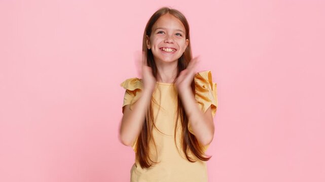Young child girl raises hands with a victory win gesture, smiling brightly and celebrating a joyful success moment. School kid isolated on pink background, full of cheerful energy and positive emotion