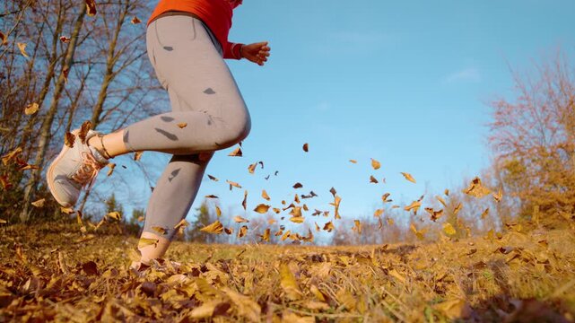 CLOSE UP, LOW ANGLE, SLOW MOTION: Sporty woman runs through windblown autumn leaves. Her sneakers and legs are lit by warm sunshine and dry foliage flies in the air. Outdoor exercise in fall season.