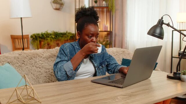 African American sleepy woman sits at table on home sofa drinks coffee tiredly after sleepless night trying to energize herself. Black girl prepares to start remote workday using laptop stay focused