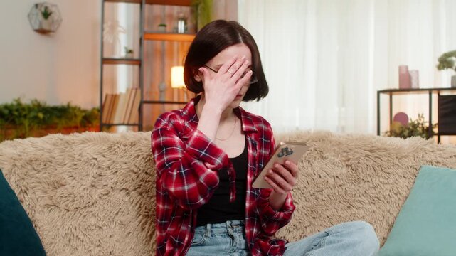 Woman looks at smartphone on home sofa with sad face after losing online lottery or failing a test. Girl reacts with visible disappointment, feeling emotional pressure and frustration from bad result