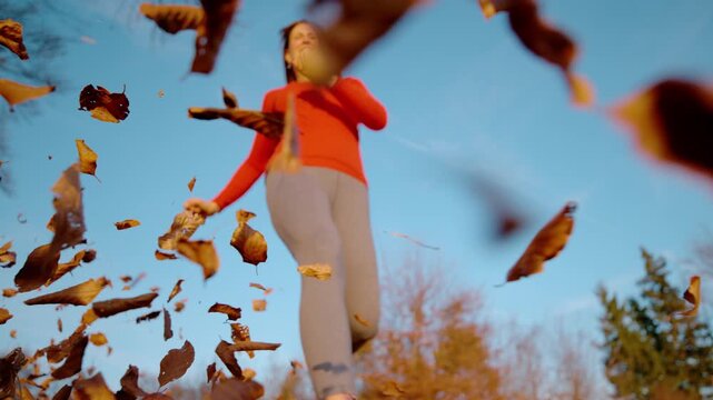 CLOSE UP, DOF, LOW ANGLE, SLOW MOTION: Smiling woman jumping over the camera as dry autumn leaves swirl through the air. Playful and energetic outdoor exercise in the sunny countryside in fall season.