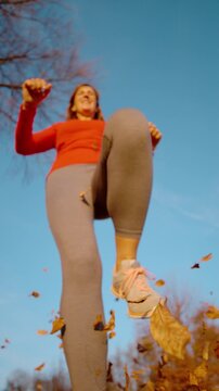 CLOSE UP, DOF, LOW ANGLE, SLOW MOTION Cheerful woman in sportswear jumps above the camera while windblown autumn leaves fly around her. Joy, movement and playful outdoor activity in rural fall setting