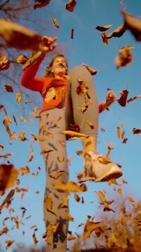 CLOSE UP, DOF, LOW ANGLE, SLOW MOTION: Smiling woman leaps over the lens as burst of windblown autumn leaves fills the frame. Flying foliage, blue sky and athletic motion during lively fall recreation