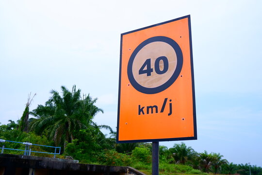 a horizontal low-angle shot capturing a bright orange 40 km/h speed limit road sign set against oil palm trees and a bright sky.