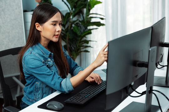 Working young beautiful Asian woman rechecking business report on pc screens comparison, wearing casual jeans shirt, analyzing sales target report business market graph at modern office. Stratagem.
