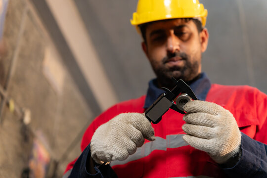 Low angle shot of technician wearing hardhat using digital vernier caliper to measure metal component in factory.