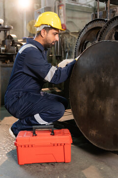 Technician wearing hardhat kneeling to inspect and repair machinery gears in industrial factory.