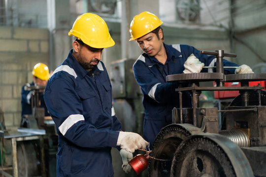 Engineers wearing hardhats working together to lubricate and repair machinery gears in industrial factory.